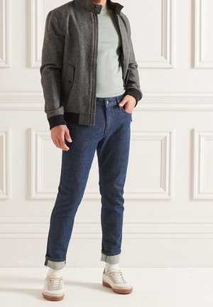 Graue Bomberjacke mit Textur und schwarzen Bündchen, kombiniert mit einem hellgrünen Hemd, blauen Jeans und weißen Sneakern, vor einem neutralen Hintergrund.