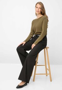 Olivengroene geribbelde gebreide trui met lange mouwen, gecombineerd met zwarte, hoge taille, brede broekspijpen en zwarte ballerina's, zittend op een houten kruk.