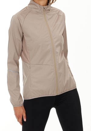 Veste légère beige à fermeture éclair, manches longues et col montant, portée avec des leggings noirs.