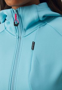 Svetlo modrá mikina na zips z textúrovanej látky, s kapucňou, ružovým ťahákom zipsu a malým čiernym logom na hrudi.