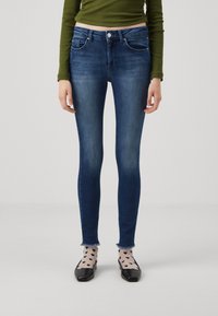 Smaljeans i mørk blå denim med en falmet vask, med midjebånd i middels høyde, fem lommer og frynsete kanter. Kombinert med svarte flate sko og mønstrede sokker.