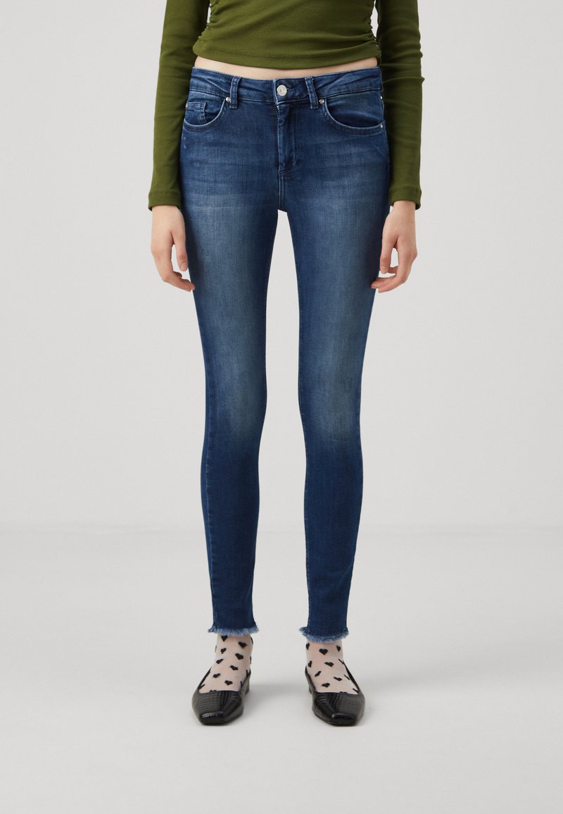 Smaljeans i mørk blå denim med en falmet vask, med midjebånd i middels høyde, fem lommer og frynsete kanter. Kombinert med svarte flate sko og mønstrede sokker.