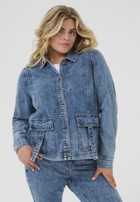 Chaqueta de mezclilla en azul claro con cierre de botones, cuello corto, dobles bolsillos en el pecho y hebillas laterales. Presenta un ajuste ligeramente relajado.