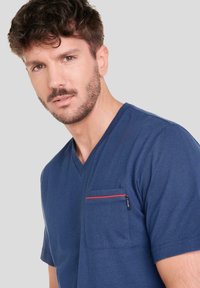 Camiseta de cuello en V azul marino hecha de tela suave, con un bolsillo en el pecho con ribete rojo y una pequeña etiqueta de la marca en el costado.