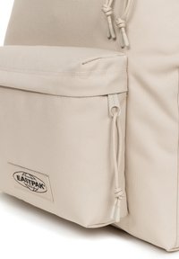 Mochila Eastpak beige con tejido texturizado, bolsillo frontal, cierre con cordón y parche con el logo. Presenta un diseño minimalista y un color neutro.