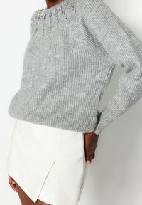 Grauer Strickpullover mit gerippter Textur und verziertem Ausschnitt. Kombiniert mit einem weißen Minirock mit hohem Schlitz.