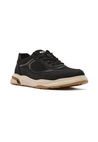 Zapatilla deportiva negra con parte superior de malla y sintético, suela marrón, puntera redondeada y detalles texturizados a lo largo de los costados.
