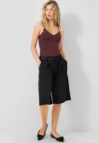 Burgundowy top na ramiączkach w połączeniu z czarnymi, plisowanymi culottami z wysokim stanem, prezentowany z rękami w kieszeniach oraz czarnymi płaskimi butami z kokardkami.