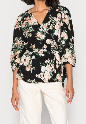 Vrouw draagt een zwarte overslagblouse met perzikkleurige en witte bloemenprint en driekwart mouwen, gecombineerd met witte broek met hoge taille.