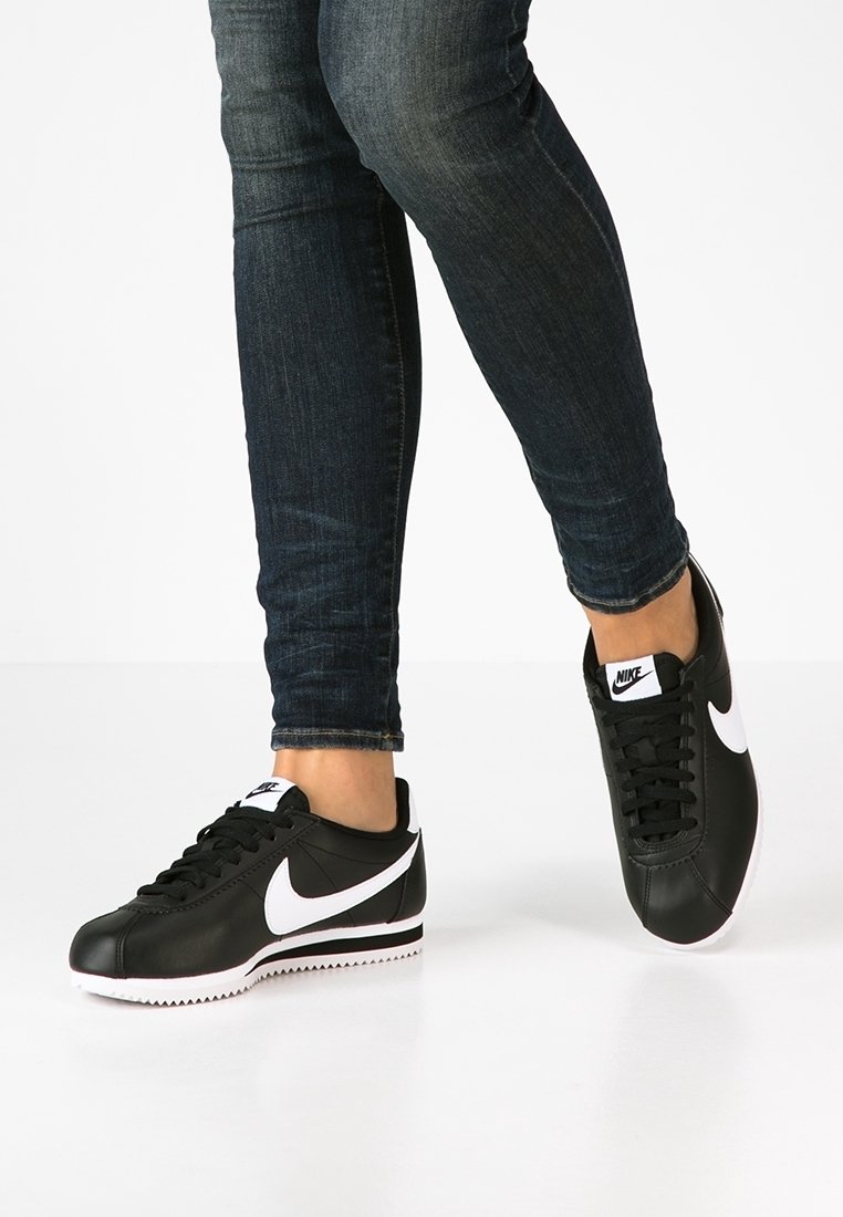 Personne portant des baskets Nike noires avec des logos swoosh blancs et un jean skinny bleu foncé, debout sur une surface blanche.