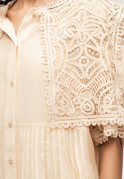 Κοντινό πλάνο ενός μπεζ πλισέ μπλουζοκαλώτσου με κουμπιά και περίπλοκο δαντελένιο κάπα με λουλουδάτα και γεωμετρικά σχέδια στον ώμο.