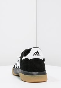 Black suede sneaker with white three-stripe design, white heel tab, and gum rubber sole. Rounded shape with padded collar.