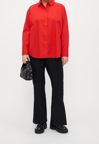 Persona che indossa una camicia rossa con bottoni, pantaloni neri a campana, scarpe platform nere con borchie e tiene in mano una borsa nera con occhielli metallici.