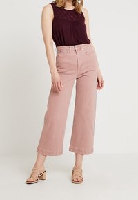 Ljusrosa vida jeans i kombination med en mörk vinröd ärmlös top med texturerade detaljer, kompletterat med neutrala sandaler med klack och remmar.