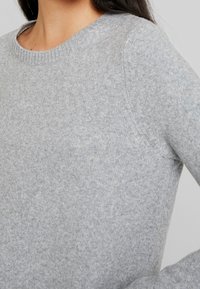 Ljusgrå tröja i mjukt stickat material. Har rundad halsringning och ribbad kant. Texturen är jämn med minimala ytdetaljer.