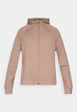 Lys brun hettejakke med glidelås, tettsittende hette, raglan-ermer og liten logo på brystet. Laget av mykt, fleksibelt stoff.