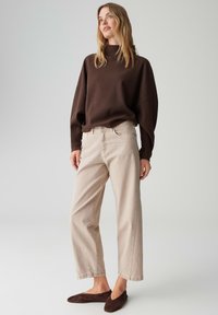 Bruine oversized trui, lichtbeige wijde jeans, en bruine platte schoenen. Zachte textuur, ontspannen pasvorm en knopen op de jeans. Neutraal kleurenpalet.