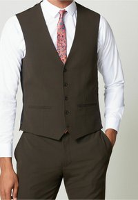 Mann in einer maßgeschneiderten dunklen Weste über einem weißen Hemd, mit einer bunten Blumen-Krawatte, die Hände in den Taschen, vor einem neutralen Hintergrund.