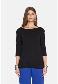 Czarny top z trzema ćwiartkami rękawów i dekoltem w łódkę, zestawiony z niebieskimi spodniami. Modelka nosi metalowe bransoletki i ma długie włosy.