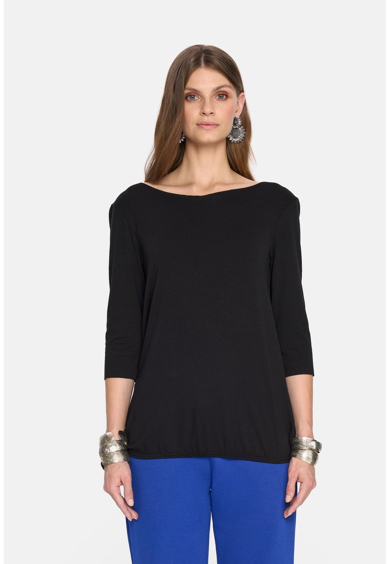 Czarny top z trzema ćwiartkami rękawów i dekoltem w łódkę, zestawiony z niebieskimi spodniami. Modelka nosi metalowe bransoletki i ma długie włosy.