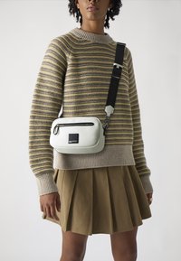 White leather crossbody bag with black strap, zip pocket, rectangular shape, smooth texture, and a logo patch in the center.