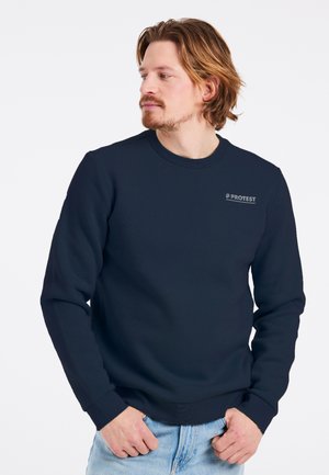 Man met lichtbruin haar en baard, gekleed in een marineblauwe trui met het logo "PROTEST", kijkt naar links, handen dichtbij de broekzakken van zijn spijkerbroek.