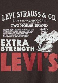 Tamsiai ruda marškinėliai su baltu ir raudonu tekstu. Pagrindinės detalės apima "LEVI STRAUSS & CO," "EKSTRA STIPRUMAS" ir piešinys suarkliu.