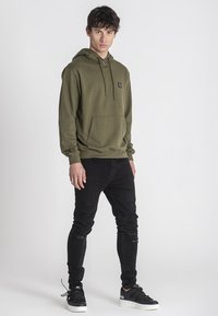 Felpa verde oliva con tasca a canguro e lacci, abbinata a jeans neri strappati e sneakers nere con suole bianche.