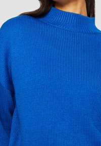 Niebieski sweter dzianinowy z wysokim kołnierzem, charakteryzujący się prążkowaną fakturą i luźnym krojem. Materiał wydaje się miękki i ciepły.