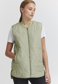 Hellgrüne, ärmellose Steppweste mit Reißverschluss, rundem Ausschnitt und seitlichen Taschen. Subtile Wellenmuster über den Stoff.