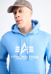 Šviesiai mėlynas džemperis su kapišonu, ant kurio balta logo su žvaigžde ir juostomis. Tekstas apima "ALPHA INDUSTRIES" ir "KNOXVILLE, TENNESSEE, JAV."