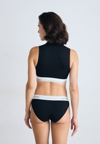 Kvinne står med ryggen til kameraet iført svart undertøysett fra Calvin Klein med hvite elastiske midjebånd mot en enkel lys bakgrunn.