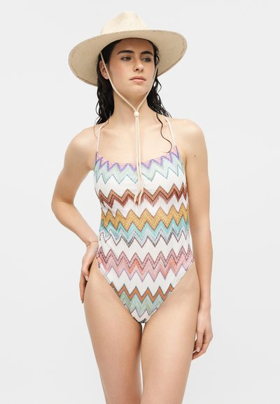 Γυναίκα με μακριά σκούρα μαλλιά που φοράει ένα καπέλο με ευρύ γείσο μπεζ χρώματος και μια πολύχρωμη ολόσωμη φόρμα μπάνιου με ζιγκ-ζαγκ μοτίβο, στέκεται μπροστά σε ένα απλό φόντο.