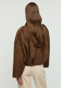 Giacca marrone in finta pelle scamosciata con maniche oversize, dettagli sulle spalle e vestibilità rilassata. Indossata sopra pantaloni beige chiaro.