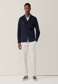 Giacca blu navy con due tasche sul petto, chiusura a bottone e maniche lunghe. Abbinata a pantaloni avorio e sneakers beige, in piedi su uno sfondo chiaro.