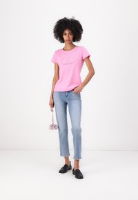 T-shirt di cotone rosa con collo rotondo e design grafico del logo, abbinata a jeans a gamba dritta blu chiaro e mocassini neri. Tiene una piccola borsa viola.