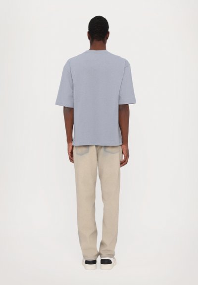 Gray cotton t-shirt with short sleeves, relaxed fit, worn with light beige pants, and white sneakers with black accents, viewed from the back.