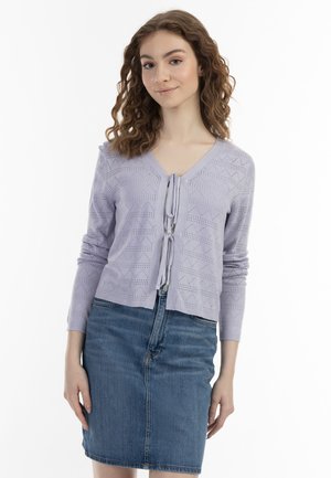 Junge Frau mit lockigem Haar trägt eine hellviolette Strickjacke mit Bindebändern und einen blauen Jeansrock, die vor einem weißen Hintergrund steht.