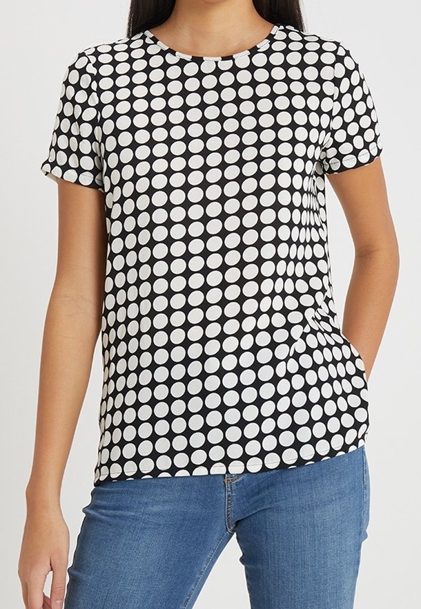 Femme portant une chemise à manches courtes à pois noirs et blancs et un jean bleu avec une main dans sa poche.
