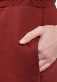 Bordowe bawełniane dżogery z prążkowanym pasem i boczną kieszenią, z ręką częściowo włożoną, ukazującą gładką fakturę i szwy.