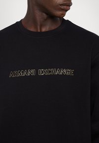 Homme portant un t-shirt à col rond noir avec l'inscription "ARMANI EXCHANGE" en lettres dorées contours, regardant vers la droite.