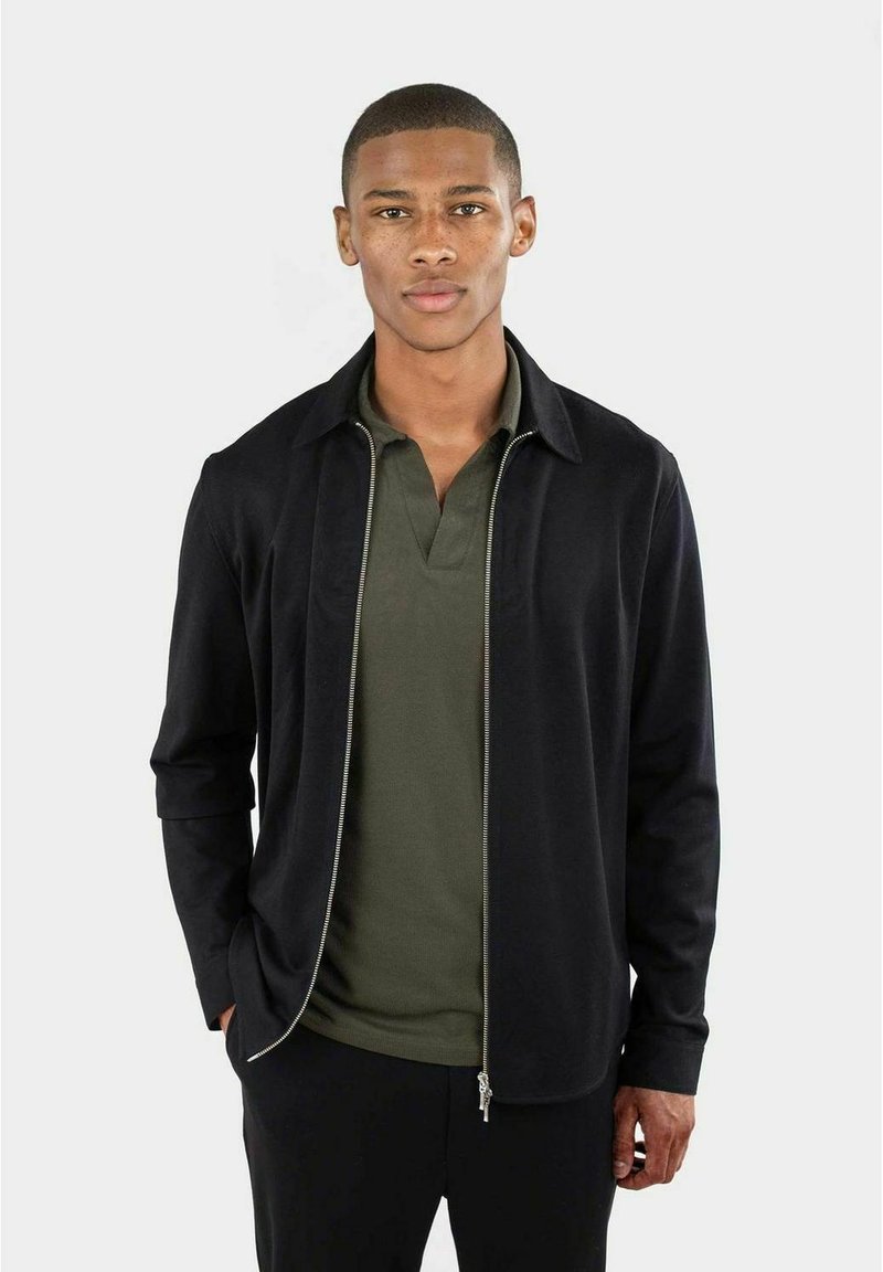Schwarze Zip-Jacke über einem olivgrünen Poloshirt, das eine glatte Textur, einen Kragen und ein tailliertes Design aufweist. Vor einem neutralen Hintergrund platziert.