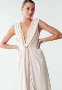 Bezrokavna, svetlo roza obleka z globokim V-izrezom in teksturirano listnato vzorcem; na pasu z naborkom in vozlom.