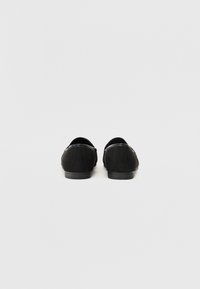 Schwarze Wildleder-Ballerinas mit niedrigem Absatz, abgerundetem Zeh und minimaler Naht. Glänzende schwarze Akzente betonen die Kante. Sauberes, schlichtes Design.
