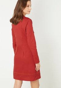 Vestido largo rojo de manga larga, confeccionado en tela suave, con forma ajustada, detalles de costura sutile en las mangas y un corte recto con pliegue en la parte posterior.