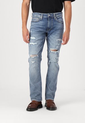 Lichtblauwe denim jeans met verwerkte details, voorzien van scheuren bij de knieën en een rechte pijp, gedragen met bruine schoenen.