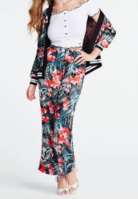 Ensemble veste et pantalon à imprimé floral noir, avec des motifs tropicaux rouges et bleu sarcelle, accompagné d'un haut blanc côtelé à épaules dénudées et de sandales blanches.
