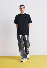 Svart kortermet t-skjorte med hvitt logo, kombinert med mønstrede grønne, grå og hvite avslappede bukser og hvite sneakers.
