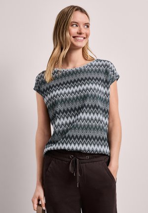 Kobieta uśmiechnięta, ubrana w szary i czarny top w zygzakowaty wzór z krótkim rękawem, zestawiony z ciemnymi spodniami ze sznurkiem, stojąca na jednolitym tle.