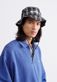 Chapéu de balde em xadrez preto e branco com cordão, usado com um pulôver azul com gola com zíper e um logótipo minimalista. Fundo liso.